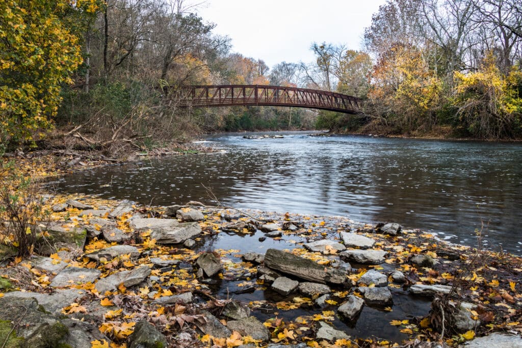 A fall landscape of the rocky shore of the Tulpehocken Creek in Reading, PA with fall foliage and a red footbridge in the background